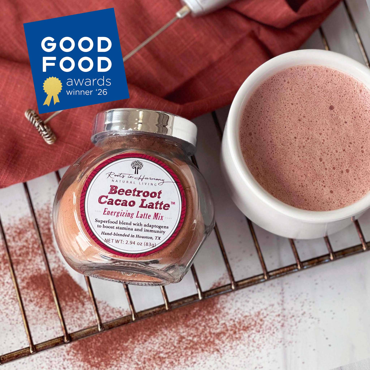 A jar of Beetroot Cacao Latte mix from Roots in the Woods Apothecary shown next to a white cup filled with frothy pink latte, on a metal cooling rack with cocoa powder scattered around, a red cloth, and a blue Good Food Awards badge.