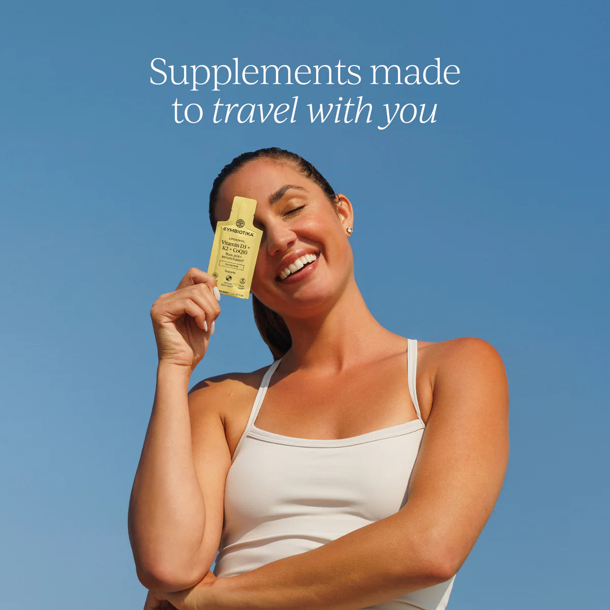 Cymbiotika Liposomal Vitamin D3 K2 CoQ10 lifestyle ad - smiling woman holding yellow supplement bottle against blue sky with text Supplements made to travel with you