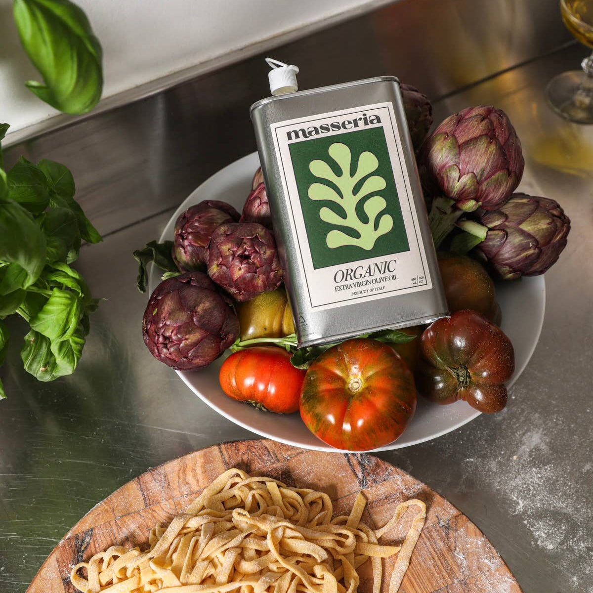 A metal tin of Masseria Organic Extra Virgin Olive Oil placed on a white plate surrounded by artichokes, tomatoes, and basil, with a wooden board of pasta in the foreground on a stainless steel countertop.
