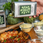 Person holding a Masseria Organic olive oil bottle over a pizza with ingredients on a wooden board.