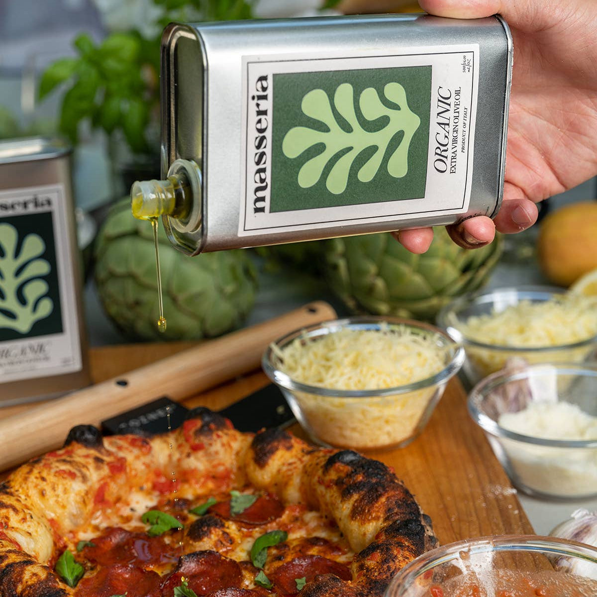Person holding a Masseria Organic olive oil bottle over a pizza with ingredients on a wooden board.