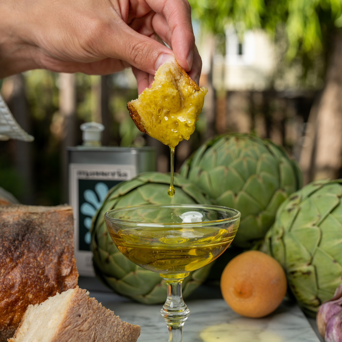 A close-up lifestyle shot of a hand dipping a piece of bread into a glass of golden olive oil, with fresh artichokes, a loaf of bread, garlic, and an apricot arranged on a table outdoors.
