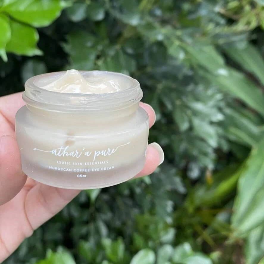 Hand holding a jar of 'athar'a pure' eye cream against a green leafy background