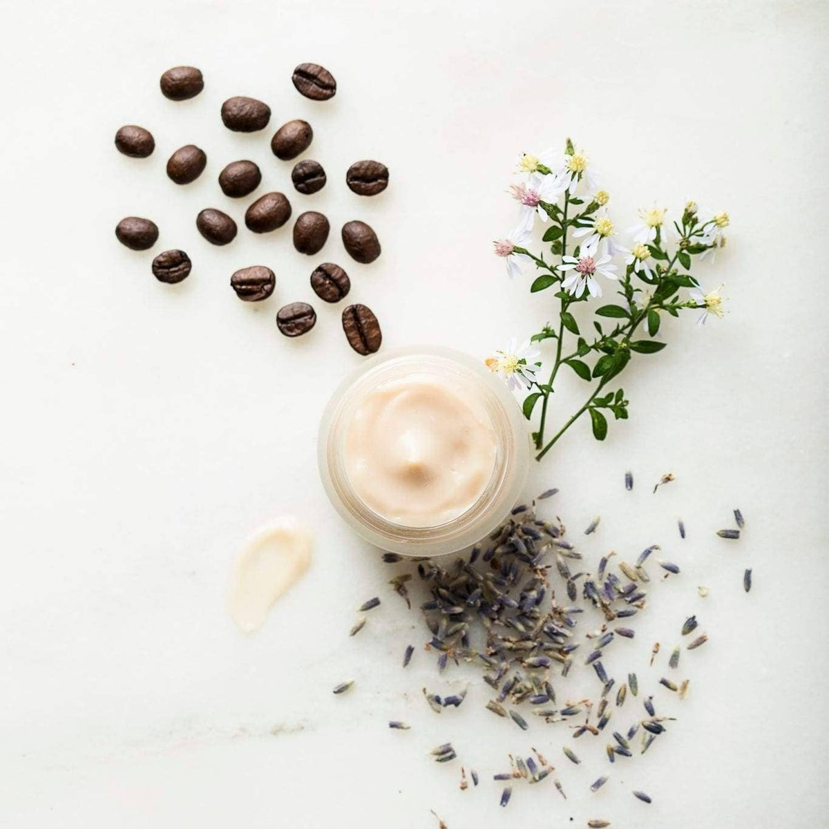 A cosmetic cream jar with a swirled beige product on top, styled on a white marble surface with scattered coffee beans, dried lavender, and small white flowers.