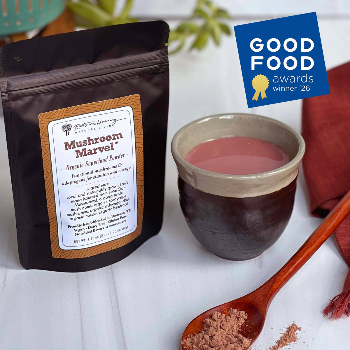A black stand-up pouch of Mushroom Marvel Organic Supplement Powder is shown next to a ceramic cup filled with pinkish-brown powder, a wooden spoon with loose powder, and a blue Good Food Awards winner badge on a white surface.
