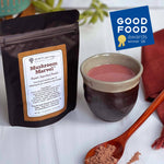 A black stand-up pouch of Mushroom Marvel Organic Supplement Powder is shown next to a ceramic cup filled with pinkish-brown powder, a wooden spoon with loose powder, and a blue Good Food Awards winner badge on a white surface.