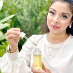 A woman in a white blouse holds a small glass bottle of pale yellow liquid and lifts a dropper toward the open bottle, with a blurred green outdoor background.