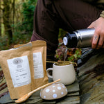 Person pouring coffee from a thermos into a mug with coffee beans and a spoon on a stone surface in a forest setting.