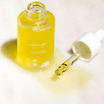 A clear glass bottle filled with golden-yellow serum, shown with its white dropper cap beside it on a light marble surface