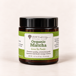 A jar of Roots in Harmony Natural Living Organic Matcha Green Tea Powder, in an amber glass jar with a black lid and a green and white label, set against a plain off-white background.