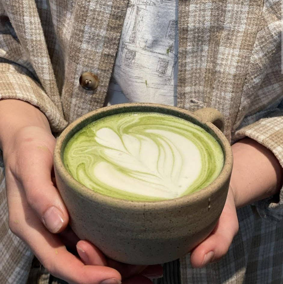 A person holding a ceramic cup of matcha latte with leaf art foam, wearing a beige plaid button-up shirt.
