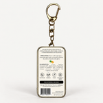 A square metal keychain container shown from the back, featuring a gold-tone keyring and clasp, with printed product information including ingredients, direction, icons, and a barcode on a white background.