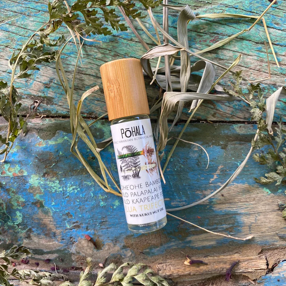 Pohala Lua Trifecta Oil roll-on bottle with bamboo cap surrounded by dried botanical plants on a rustic blue-painted wooden surface