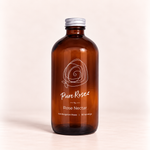 An amber glass bottle of Pure Rose Rose Nectar with a silver metal cap, set against a clean, light beige background.