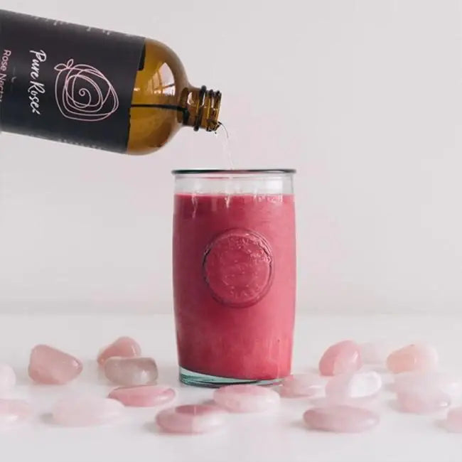 Bottle of 'Pure Rose' rose water being poured into a glass with pink stones and a white background.