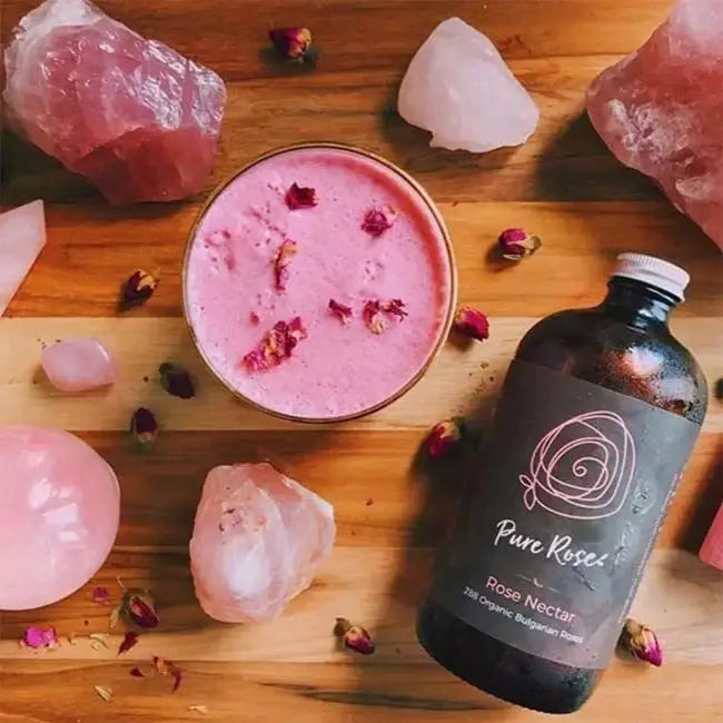 Pink rose petal-infused drink in a glass with a bottle of Pure Rose rose nectar on a wooden surface with pink rose petals and crystals.