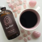 Bottle of Pure Rose' Rose Nectar next to a cup of coffee and pink stones on a light surface