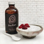 Bottle of Pure Rose' rose nectar next to a bowl of dessert with raspberries on a white background.