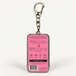 A rectangular keychain with a pink back panel showing product instructions, ingredients, and a barcode, attached to a gold-tone metal key ring and clasp, on a plain white background.